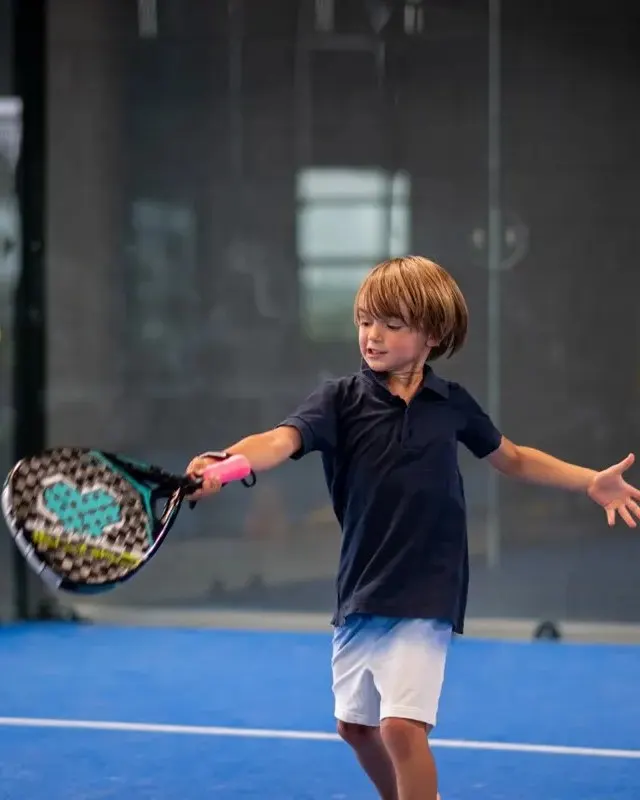 A young pupil plays a forehand while a coach feeds balls on an indoor court